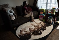 A photo of a Black woman sitting for a portrait indoors by a window. The carpet below her couch has an image of four young Black girls praying.