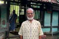 A portrait of former smallpox eradication worker Shahidul Haq Khan. He wears glasses, and a short, gray beard covers his jawline. He stands in front of his home, which has bright blue walls with dark wooden support beams.