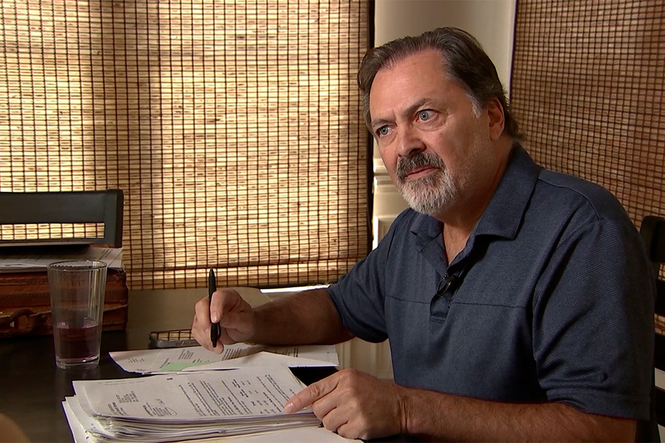 A photo of a man sitting with a pen and paperwork, looking at an interviewer out of frame.