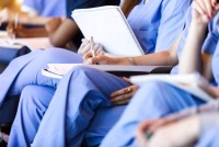 A diverse group of unrecognizable medical practitioners wearing scrubs listen to a lecture and take notes.