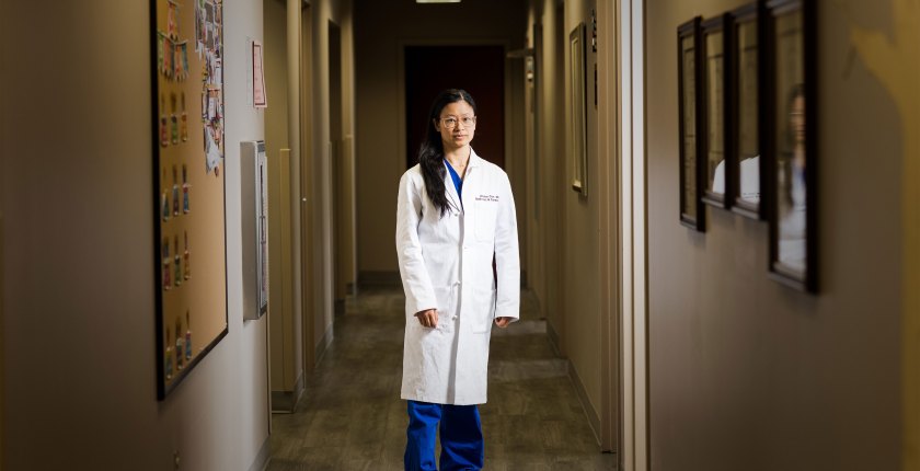 A woman with long dark hair and wearing a white doctor's coat stands in a hallway with arms by her side and looks at the camera.